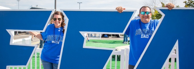 A UB Mom and UB Dad stand proudly behind large wooden letters reading "BUFFALO" in coordinating royal blue UB MOM and UB DAD shirts. 